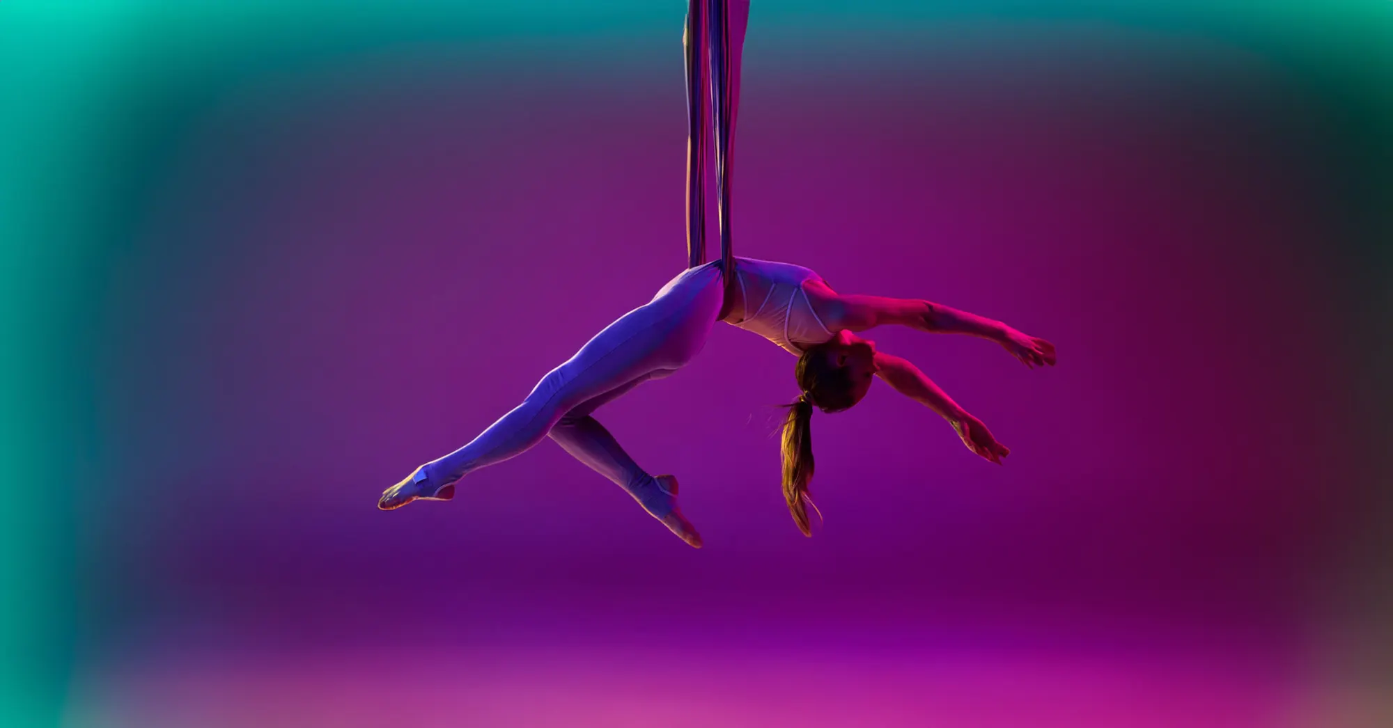 <p>Young flexible girl lying back, doing aerial fly yoga training in a purple and turquoise lit-up studio.</p>