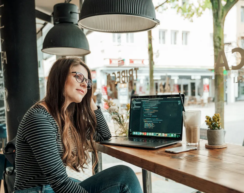 <p>A woman sitting by a window at a café with her laptop and a glass of coffee, coding.</p>