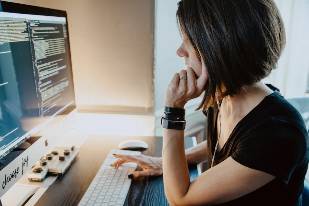 <p>A woman sitting by a desk, in front of a computer, coding.</p>